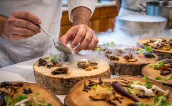 Un chef dresse avec art un plat gastronomique avec des champignons et des légumes verts sur des tranches de bois.